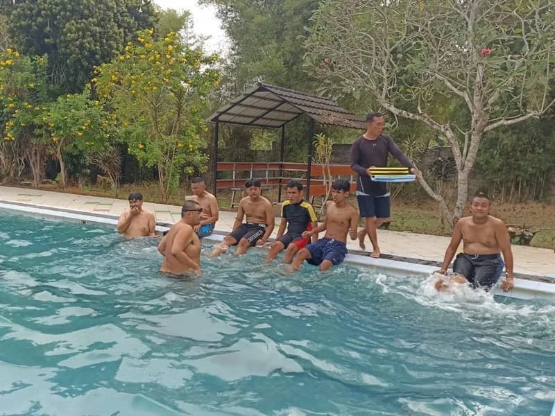 Siswa duduk berjajar di tepi kolam sambil menggerakkan kaki di air, seorang pendamping membawa papan apung