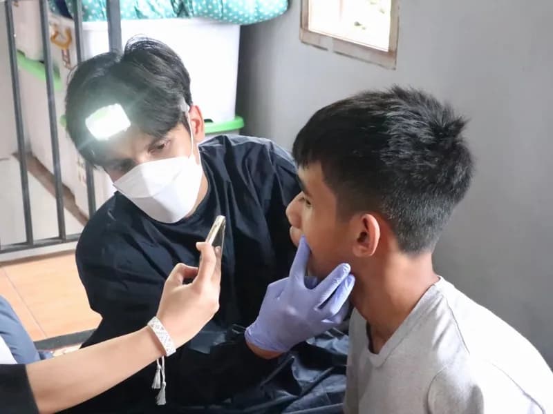 Petugas kesehatan berlampu kepala memeriksa gigi seorang siswa dengan bantuan penerangan ponsel